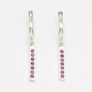 May include: A pair of silver hoop earrings with a vertical bar pendant. Each bar is adorned with a line of small, round, red gemstones. The earrings are displayed against a white background, highlighting the jewelry's design.