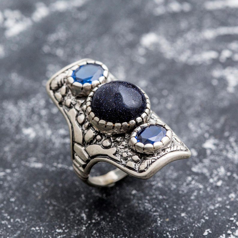 May include: A silver ring with a large blue stone and two smaller blue stones. The ring has an intricate design and is made of metal.