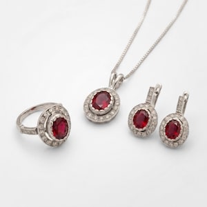 May include: A jewelry set consisting of a ring, a pair of earrings, and a pendant necklace. Each piece features a central oval-cut deep red gemstone surrounded by a halo of small white stones set in a silver-toned metal frame on a thin chain.