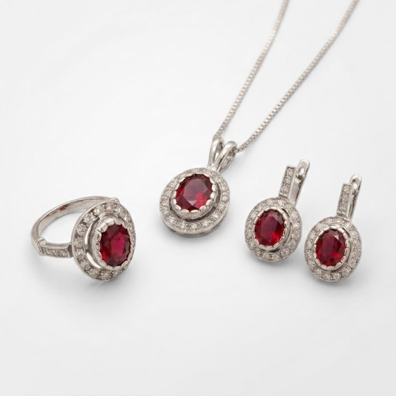 Puede incluir: Un conjunto de joyer&iacute;a compuesto por un anillo, un par de pendientes y un collar con colgante. Cada pieza presenta una piedra preciosa roja oscura de corte ovalado rodeada por un halo de peque&ntilde;as piedras blancas en un marco de metal plateado.