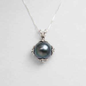 May include: A dark gray spherical pearl pendant suspended from a thin silver chain. The pearl is encased in a decorative silver setting featuring intricate leaf-like metalwork at the top and bottom, providing a secure mount for the gemstone.