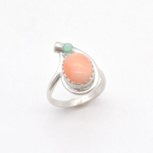 May include: A silver ring with a pink coral stone and a green gemstone. The ring has a unique design with a curved band.