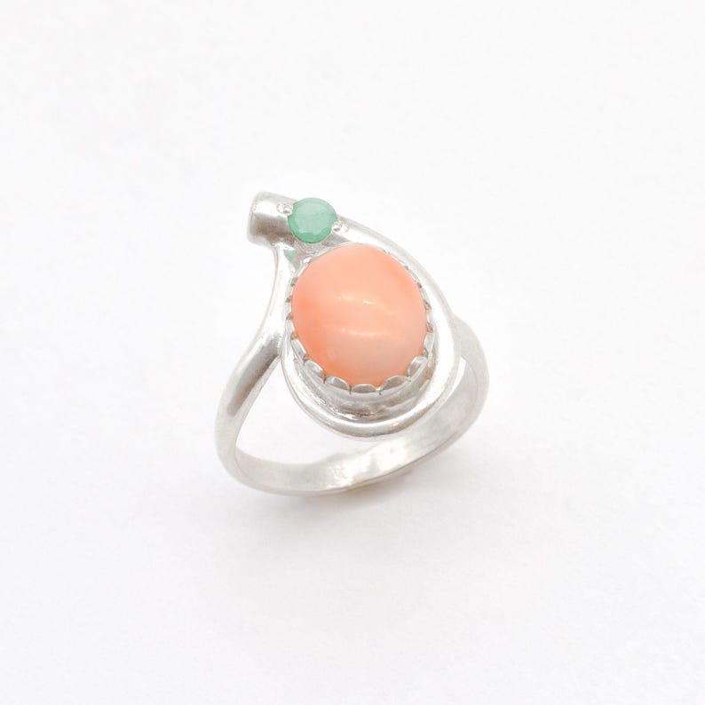 May include: A silver ring with a pink coral stone and a green gemstone. The ring has a unique design with a curved band.
