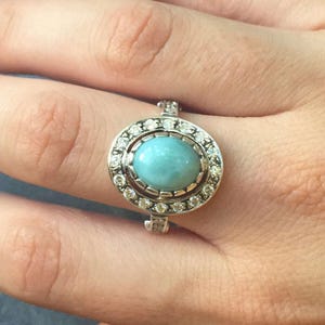 Può includere: Un anello d'argento con una grande pietra ovale azzurra, circondata da un alone di pietre più piccole e trasparenti. L'anello è mostrato su una mano, evidenziando i dettagli del design. La fascia dell'anello presenta un delicato motivo strutturato.