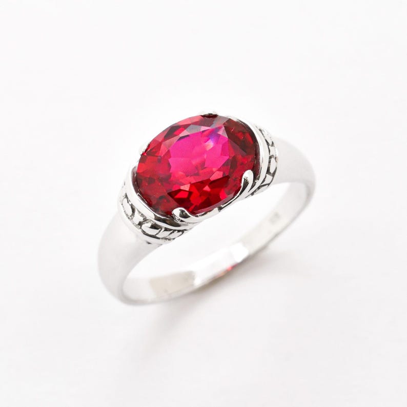 May include: A silver ring featuring a large, oval-cut, vibrant red gemstone. The gemstone is the focal point, set in a decorative silver band with ornate detailing on the sides. The ring is displayed against a white background.