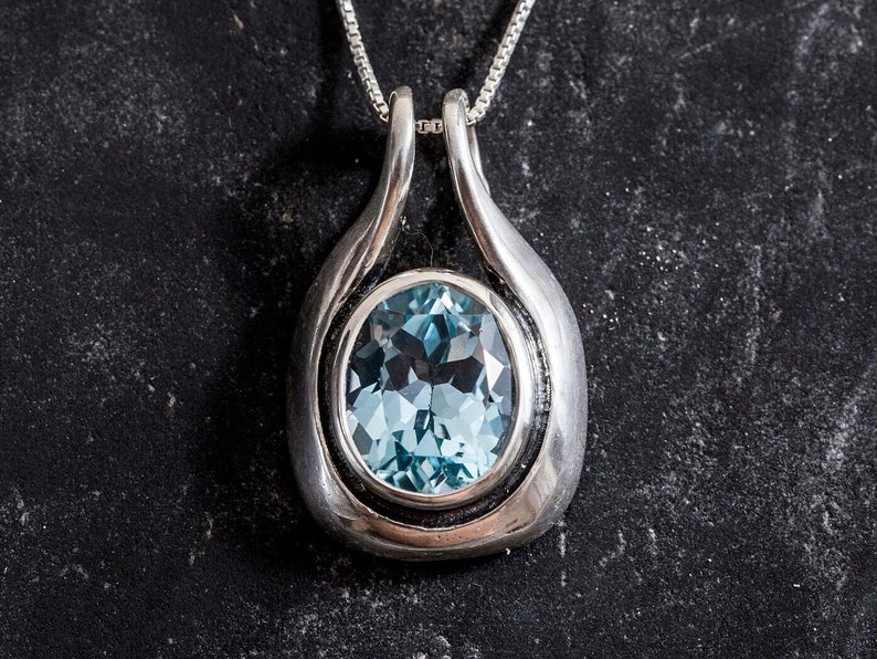 May include: A silver pendant necklace with a light blue oval gemstone set in a silver bezel. The pendant is shaped like a teardrop and has a smooth, polished finish.
