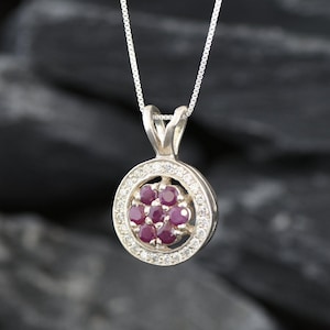 Op de afbeelding: Een zilveren ketting met een ronde hanger met een cluster van rode robijnstenen omgeven door een halo van kleinere witte stenen.
