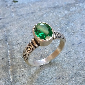 May include: A silver ring with an oval, emerald-green gemstone. The ring has an ornate, engraved design on the band and around the gemstone setting. The gemstone is the focal point, with a bright, vibrant green color.