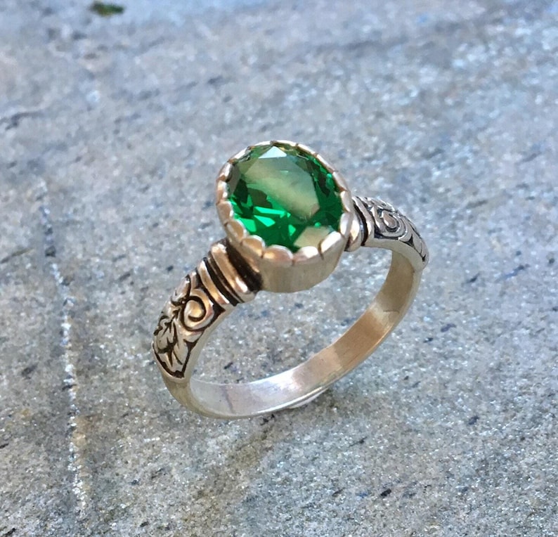 May include: A silver ring with an oval, emerald-green gemstone. The ring has an ornate, engraved design on the band and around the gemstone setting. The gemstone is the focal point, with a bright, vibrant green color.