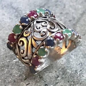 Puede incluir: Un anillo de plata con un diseño de filigrana y una banda de piedras preciosas de colores. Las piedras preciosas son rojas, verdes y azules.