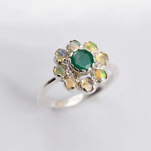May include: A silver ring with a green emerald gemstone surrounded by smaller opalescent stones, forming a flower shape.