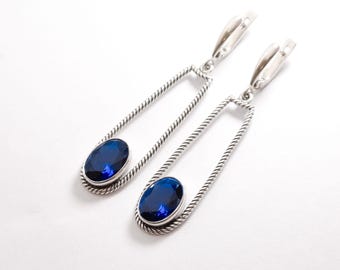 May include: A pair of silver dangle earrings with a blue oval gemstone set in each. The earrings feature a twisted rope design and a rectangular frame.