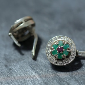 May include: Silver earrings with a floral design. Each earring features a cluster of green and red gemstones surrounded by a halo of small, clear stones. The earrings are set against a dark grey surface.