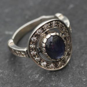 K&ouml;nnte beinhalten: Ein silberner Ring mit einem dunkelblauen Edelstein, der von einem Kreis aus kleineren, klaren Edelsteinen umgeben ist.