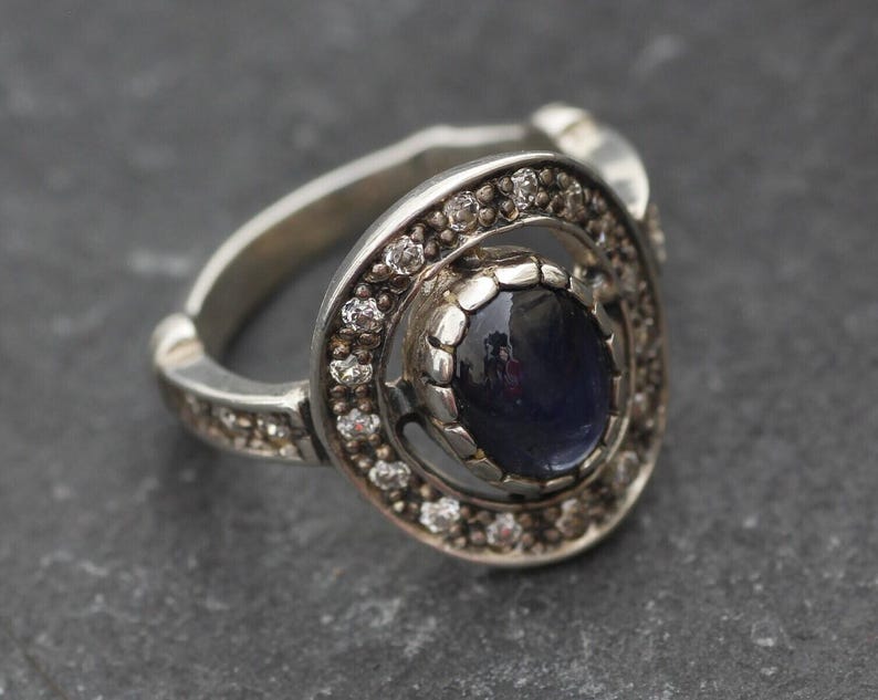K&ouml;nnte beinhalten: Ein silberner Ring mit einem dunkelblauen Edelstein, der von einem Kreis aus kleineren, klaren Edelsteinen umgeben ist.