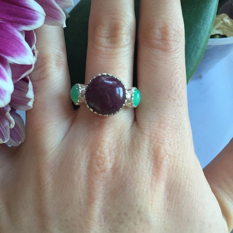 May include: A silver ring featuring a large, round, dark red gemstone flanked by two smaller, oval, green gemstones. The ring is set against a hand, with a purple and white flower in the background.