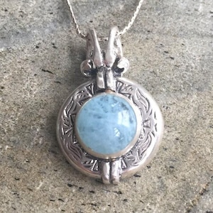 May include: A silver pendant necklace with a round, light blue aquamarine stone set in a silver frame with intricate designs. The pendant is attached to a silver chain.
