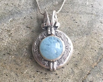 May include: A silver pendant necklace with a round, light blue aquamarine stone set in a silver frame with intricate designs. The pendant is attached to a silver chain.
