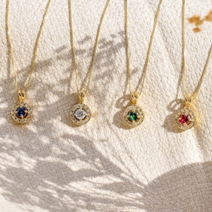 Peut inclure: Quatre colliers en or avec des pendentifs en pierres précieuses. Chaque pendentif présente une pierre centrale entourée de petites pierres. Les pierres sont bleues, claires, vertes et rouges. Les colliers sont présentés sur une surface texturée blanc cassé.