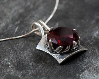 May include: A silver pendant necklace with a large, faceted, deep red gemstone. The gemstone is set in a decorative silver setting with leaf-like details. The pendant hangs from a delicate silver chain, creating a striking contrast.