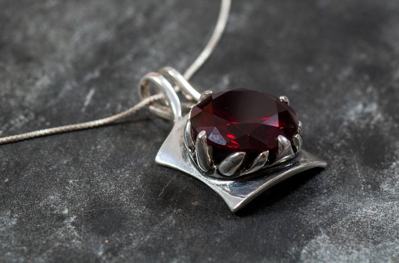 May include: A silver pendant necklace featuring a large, faceted, deep red gemstone. The gemstone is set in a decorative silver setting with leaf-like details. The pendant hangs from a delicate silver chain, creating a striking contrast.