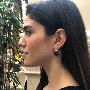 Puede incluir: Una mujer con el pelo largo y oscuro que lleva un par de pendientes de plata con una piedra de color morado oscuro.