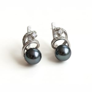 May include: A pair of silver earrings with a swirling design. Each earring has a dark grey pearl suspended below a loop decorated with small, clear gemstones. A larger, clear gemstone is set at the top of the loop.