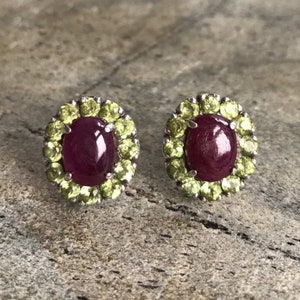 May include: A pair of oval-shaped ruby stud earrings, each surrounded by a halo of small, light green peridot stones. The earrings are set in a silver metal. The background is a textured, gray surface.