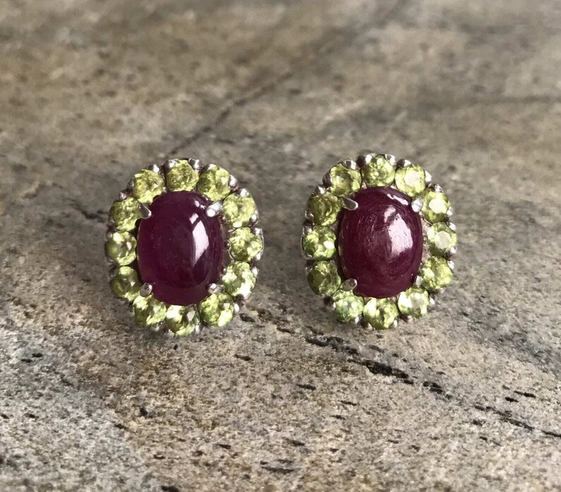 May include: A pair of oval-shaped ruby stud earrings, each surrounded by a halo of small, light green peridot stones. The earrings are set in a silver metal. The background is a textured, gray surface.