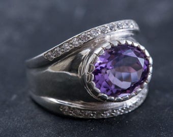 May include: A silver ring with a large oval amethyst gemstone surrounded by smaller clear gemstones. The ring has a wide band with a textured design.