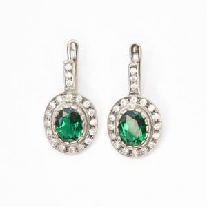 May include: A pair of silver earrings featuring a green oval gemstone set within a halo of smaller white gemstones.