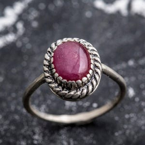 May include: A silver ring with a pink oval gemstone set in a decorative silver bezel. The ring is on a dark gray background.