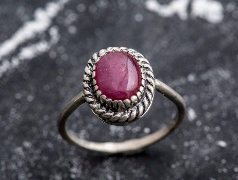 May include: A silver ring with a pink oval gemstone set in a decorative silver bezel. The ring is on a dark gray background.