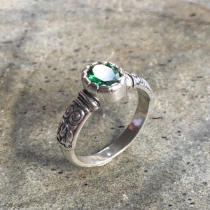 May include: A silver ring with a large, round, emerald-green gemstone. The ring has an ornate, engraved design on the band and around the gemstone setting. The ring is displayed on a textured, gray surface.
