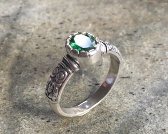 May include: A silver ring with a large, round, emerald-green gemstone. The ring has an ornate, engraved design on the band and around the gemstone setting. The ring is displayed on a textured, gray surface.