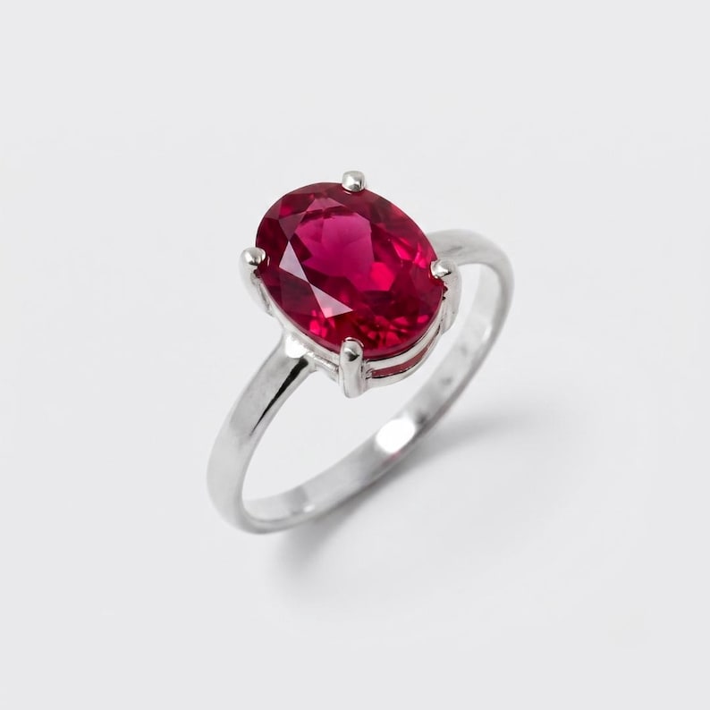 Pode incluir: Um anel de tom prateado com uma grande pedra preciosa de cor vermelha vibrante e corte oval, fixada por um engaste de quatro garras. A banda &eacute; lisa e polida, apresentando um design simples e limpo que destaca a cor vermelha intensa.