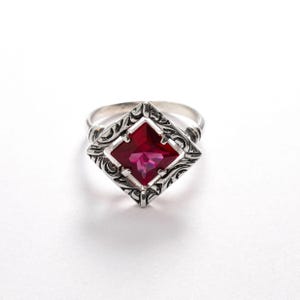 Peut inclure: Une bague en argent avec un motif orné et une pierre rouge carrée.