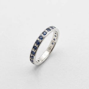 May include: A sterling silver band ring featuring a channel setting with a row of small, round, deep blue sapphires. The inner surface of the band is clearly stamped with the hallmark 925, indicating the silver content of the jewellery piece.