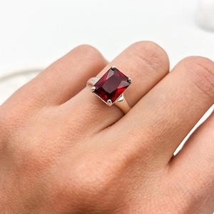 May include: A silver ring featuring a large, square-shaped red gemstone.