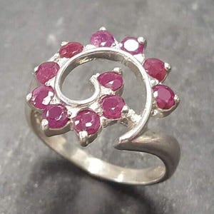 May include: A silver ring with a swirling design set with pink gemstones. The ring is open and adjustable.