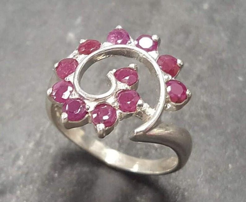 May include: A silver ring with a swirling design set with pink gemstones. The ring is open and adjustable.