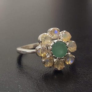 May include: A silver ring with a green gemstone surrounded by smaller white gemstones, forming a flower shape.