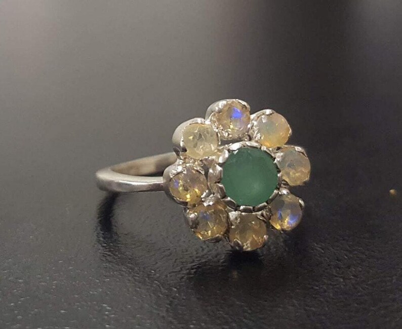 May include: A silver ring with a green gemstone surrounded by smaller white gemstones, forming a flower shape.