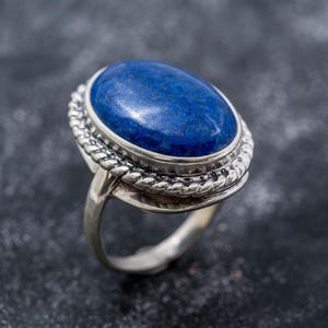 May include: A silver ring with an oval, deep blue gemstone. The gemstone is set in a silver bezel with a twisted rope design around the stone. The ring band is smooth and polished.