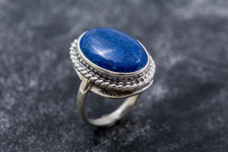 May include: A silver ring with an oval, deep blue gemstone. The gemstone is set in a silver bezel with a twisted rope design around the stone. The ring band is smooth and polished.