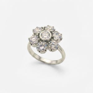 May include: A silver-toned ring featuring a cluster of seven round, clear, faceted stones arranged in a floral pattern. The center stone is bezel-set, while the six surrounding stones are prong-set, creating a circular, flower-like design.