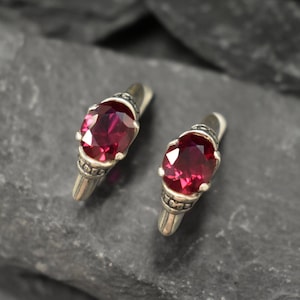 May include: A pair of silver hoop earrings with a red gemstone in each. The earrings have a detailed design with small silver beads.