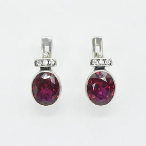 May include: A pair of silver earrings with a round, dark red gemstone and a row of small, clear gemstones on the top.