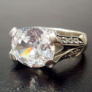 Puede incluir: Un anillo de plata con una gran piedra preciosa ovalada transparente rodeada de piedras preciosas transparentes m&aacute;s peque&ntilde;as. El anillo tiene un dise&ntilde;o delicado y ornamentado.