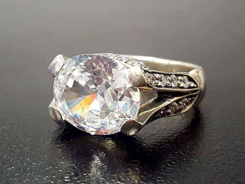 Puede incluir: Un anillo de plata con una gran piedra preciosa ovalada transparente rodeada de piedras preciosas transparentes m&aacute;s peque&ntilde;as. El anillo tiene un dise&ntilde;o delicado y ornamentado.